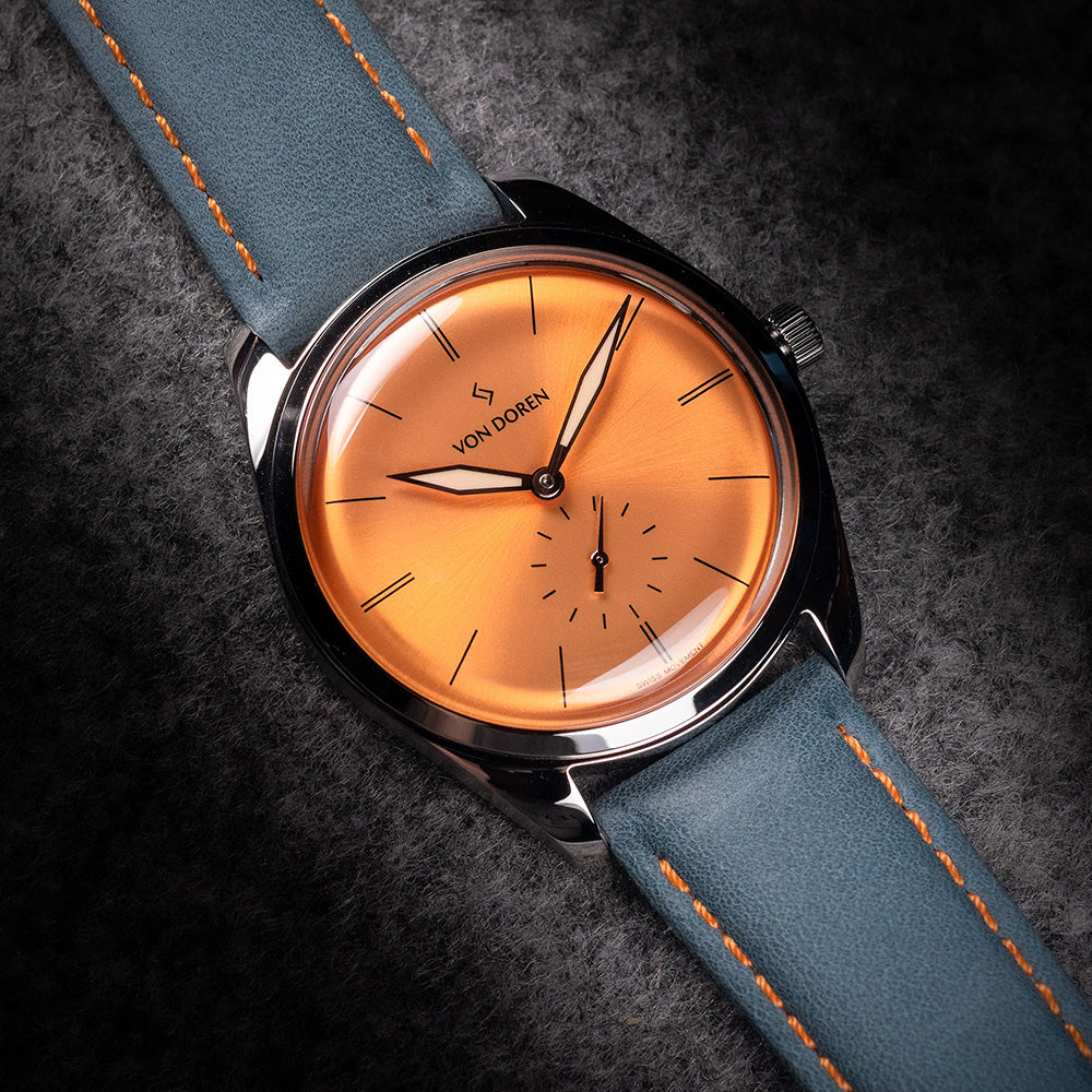 Von Doren-klokke med oransje sunburst-tallskive og blå rem med oransje søm.