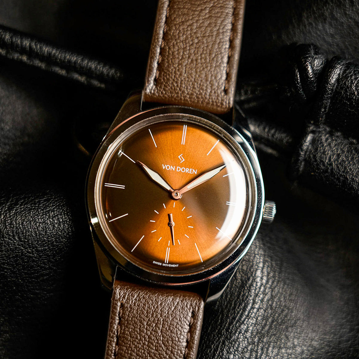 Von Doren klokke. Brun skive med brun skinnreim, liggende på sort lær.