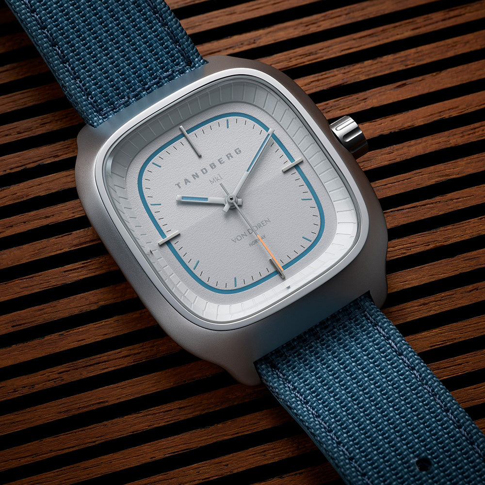 Tandberg klokke med hvit skive og blå reim liggende på