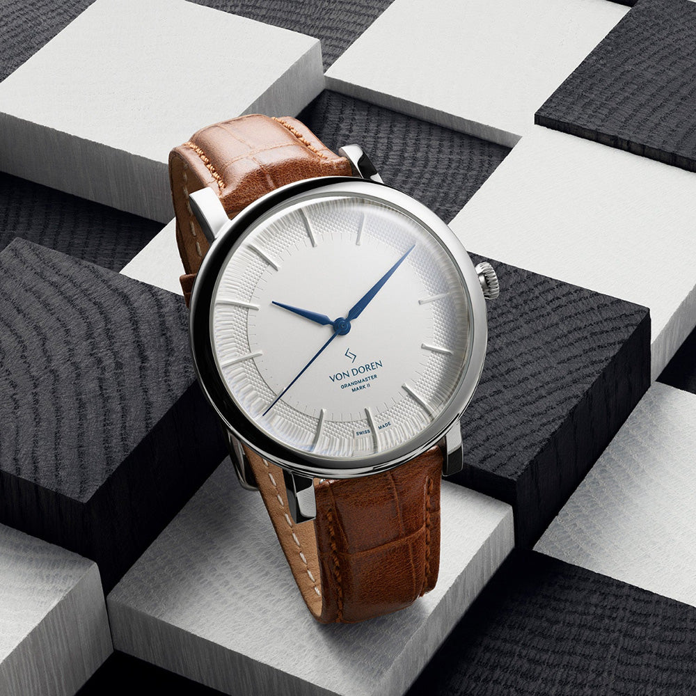 Von Doren-klokke med hvit tallskive og brun rem på sjakkmønstret bakgrunn.